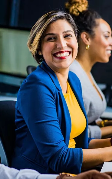 A woman in a blue jacket smiles at an office table with a colleague beside her