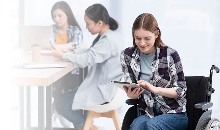 A woman using a tablet while seated in a wheelchair at a table with coworkers