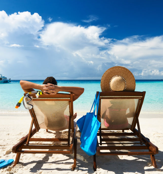 Two travelers relax on beach chairs beside a bright blue ocean.