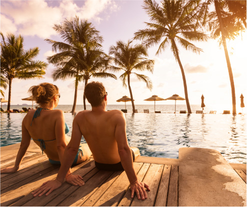 Two travelers sit beside a tropical pool at sunset, with palm trees behind them.